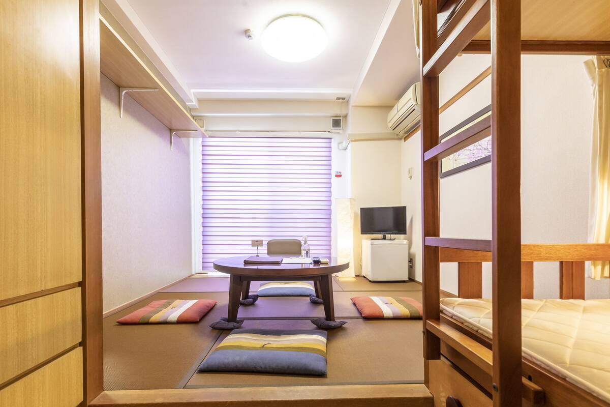 A compact room with a bunk bed, a small desk with a chair, a television, and a window with blinds.