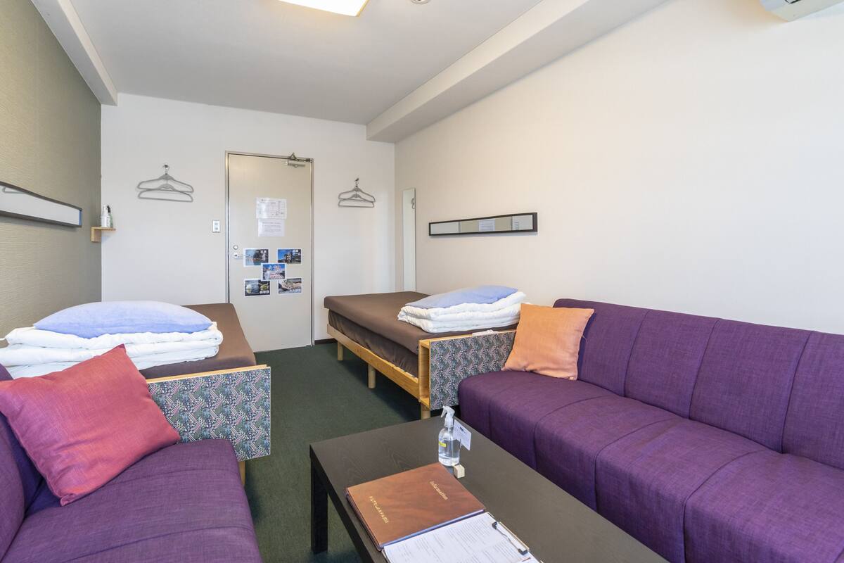 A hotel room with a bed, a sofa, a small table, and a wall-mounted air conditioner.