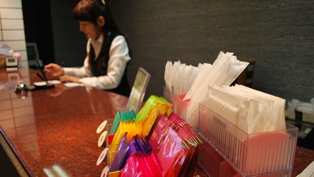 A reception desk with a woman working behind it, a colorful display of envelopes, and a clear plastic organizer with papers. - Sky Hotel Uozu Annex (Uozu)