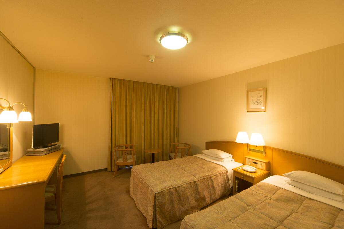 Quarto de hotel com duas camas, uma escrivaninha com televisão, uma cadeira e uma mesinha de cabeceira com luminárias.