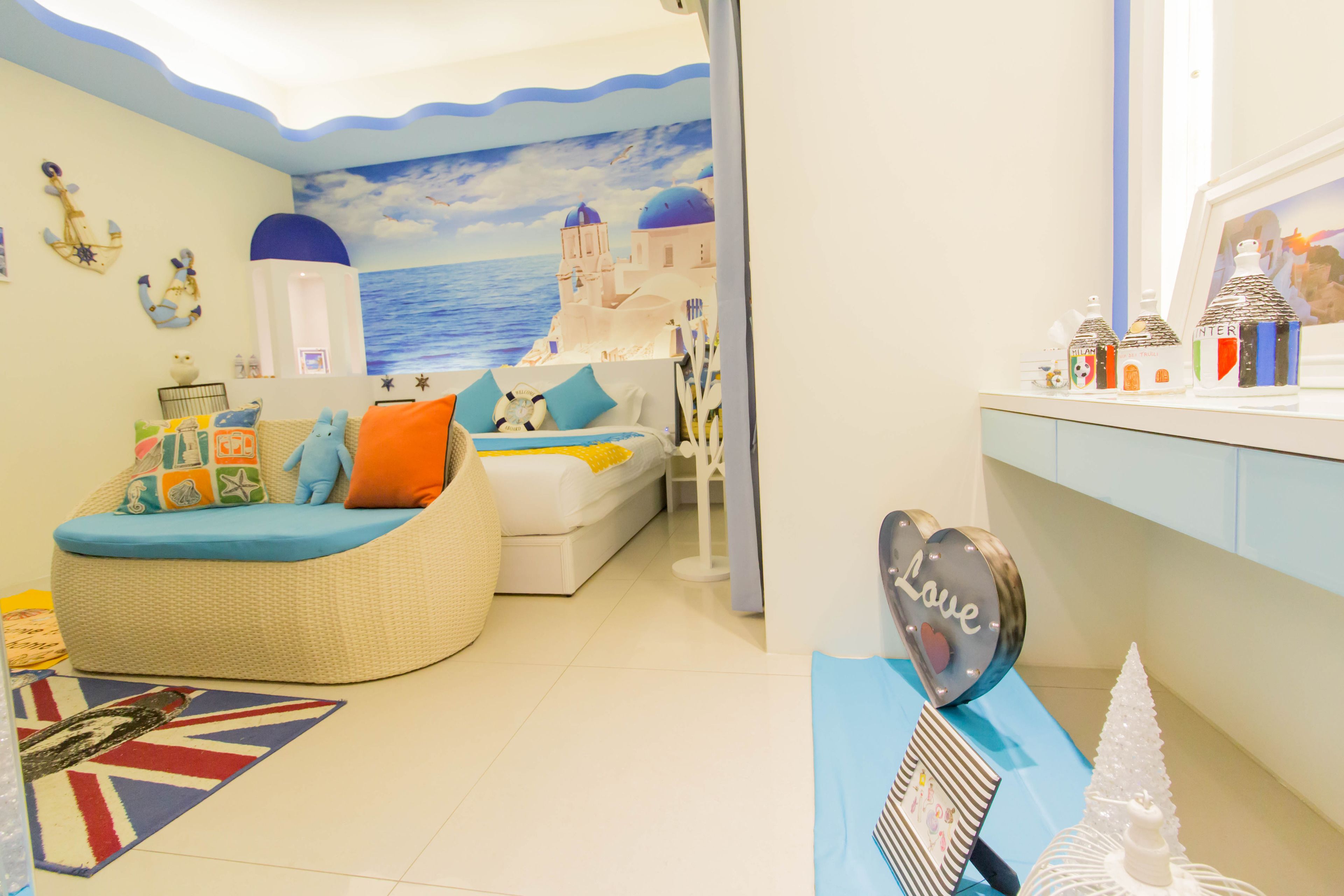 A room with a curved sofa, a bed, a small table with decorative items, and a wall mural of a coastal scene.