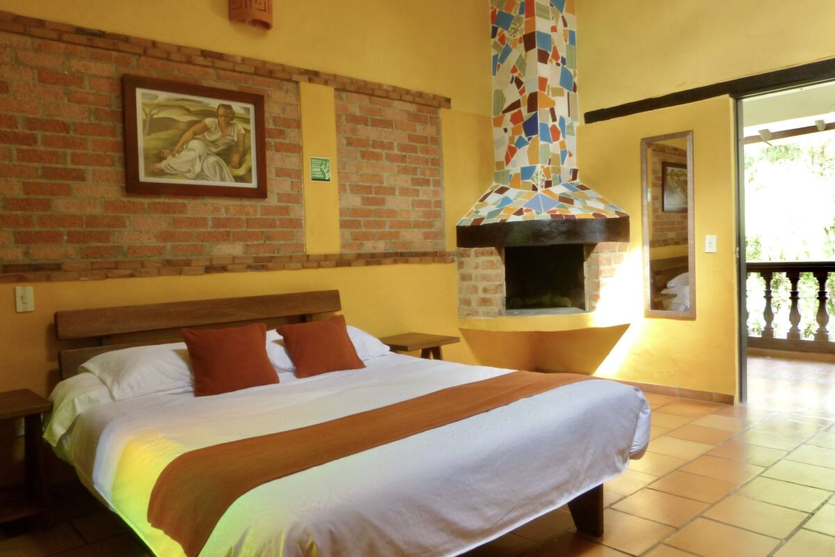 A hotel room with a brick wall, a bed with white and brown bedding, a fireplace, and a balcony with a railing.