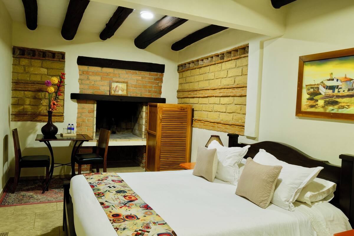 A bedroom with a bed, a desk, a chair, a fireplace, and a painting on the wall.