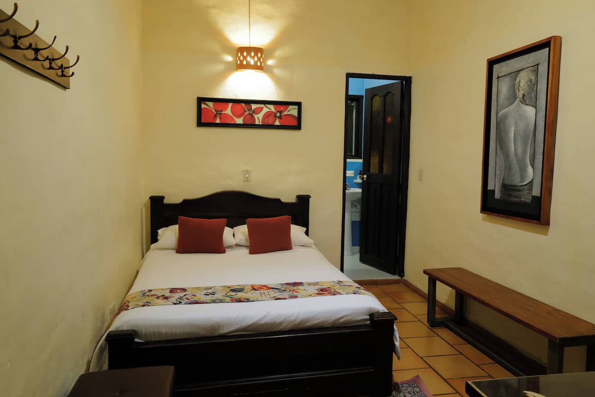 A bedroom with a bed, a bench, a painting, and a door leading to another room.