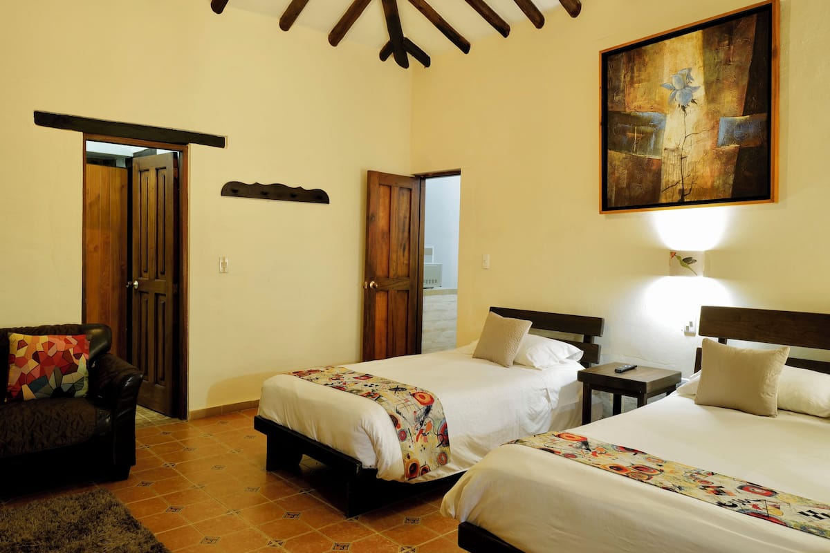 A hotel room with two beds, a sofa, a painting on the wall, and a small table with a remote control.