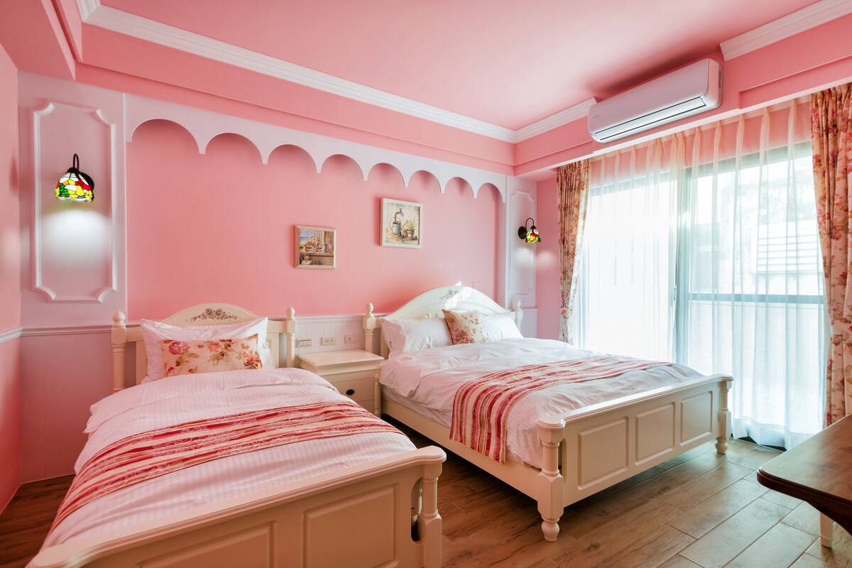 A pink-themed bedroom with two beds, a nightstand, and a window with curtains.