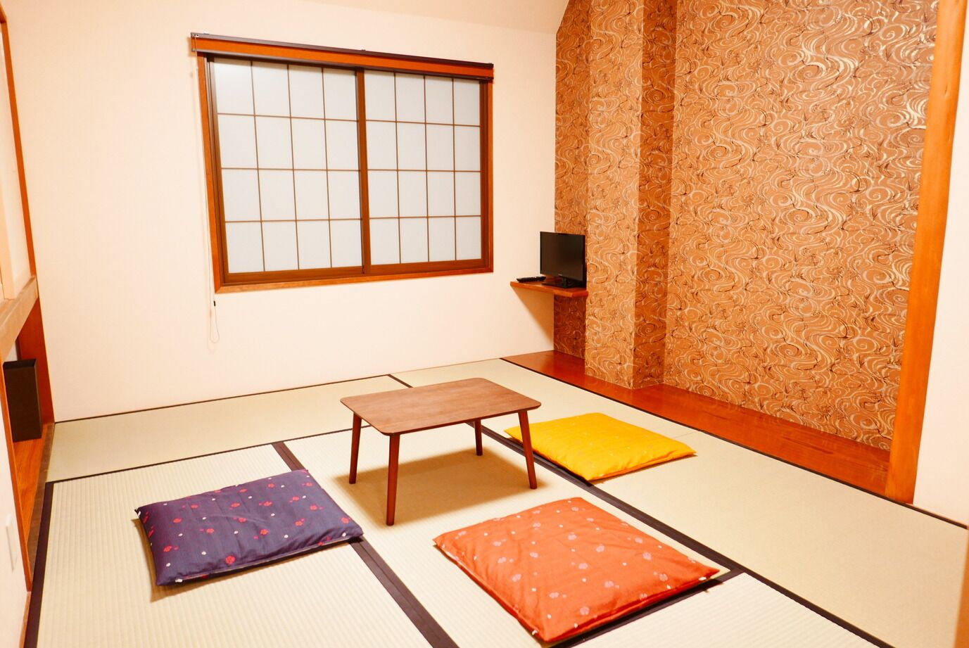 A tatami mat room with a sliding door, a small table, cushions, and a TV on a shelf.
