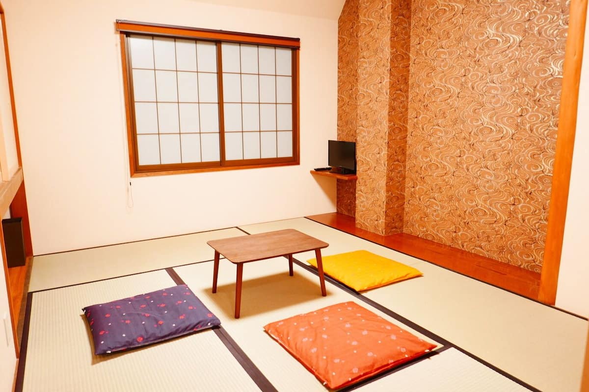A tatami mat room with a sliding door, a small table, cushions, and a TV on a shelf.