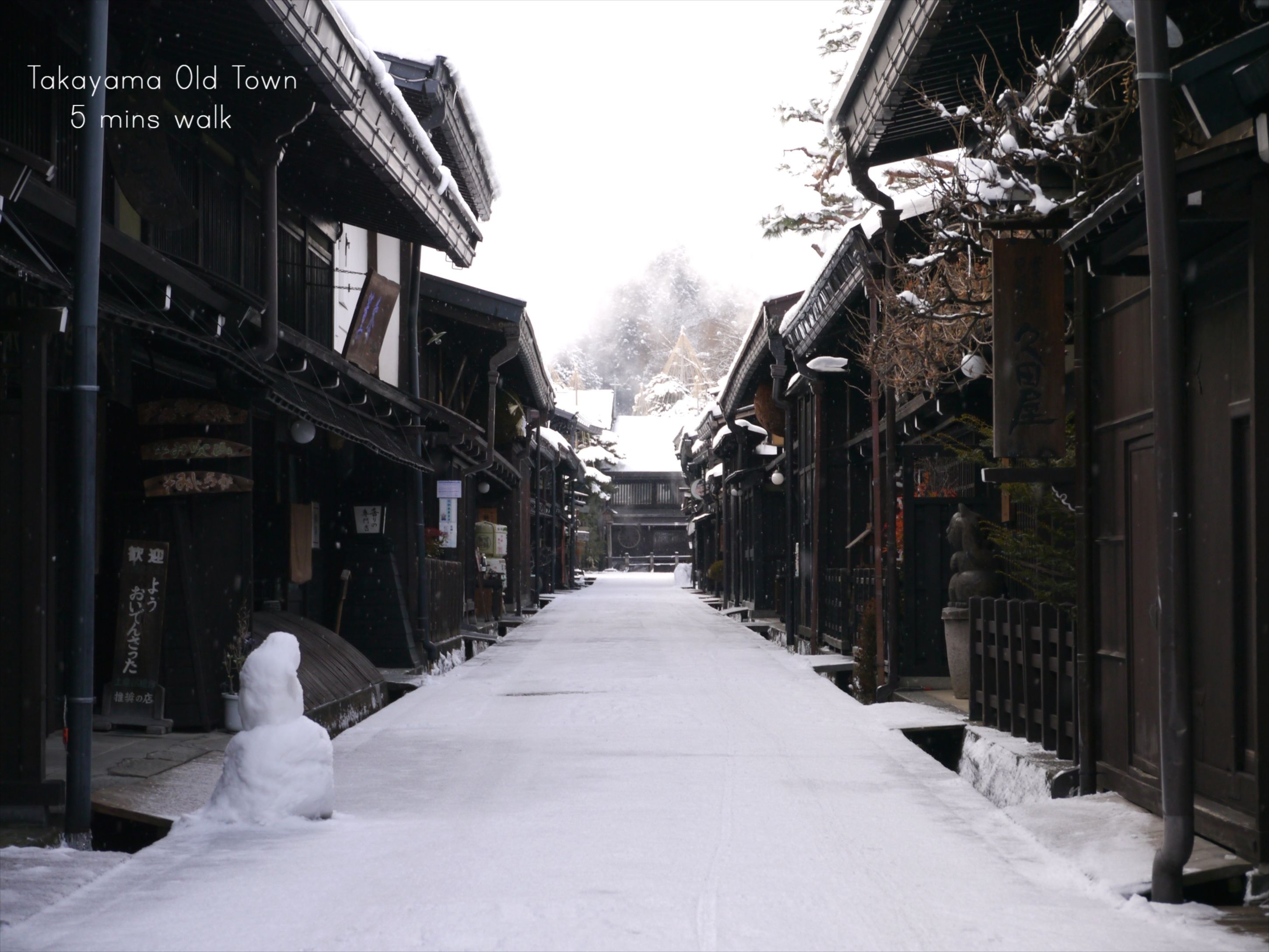 A snow-covered street in Takayama Old Town, lined with traditional wooden buildings and a snowman at the entrance.