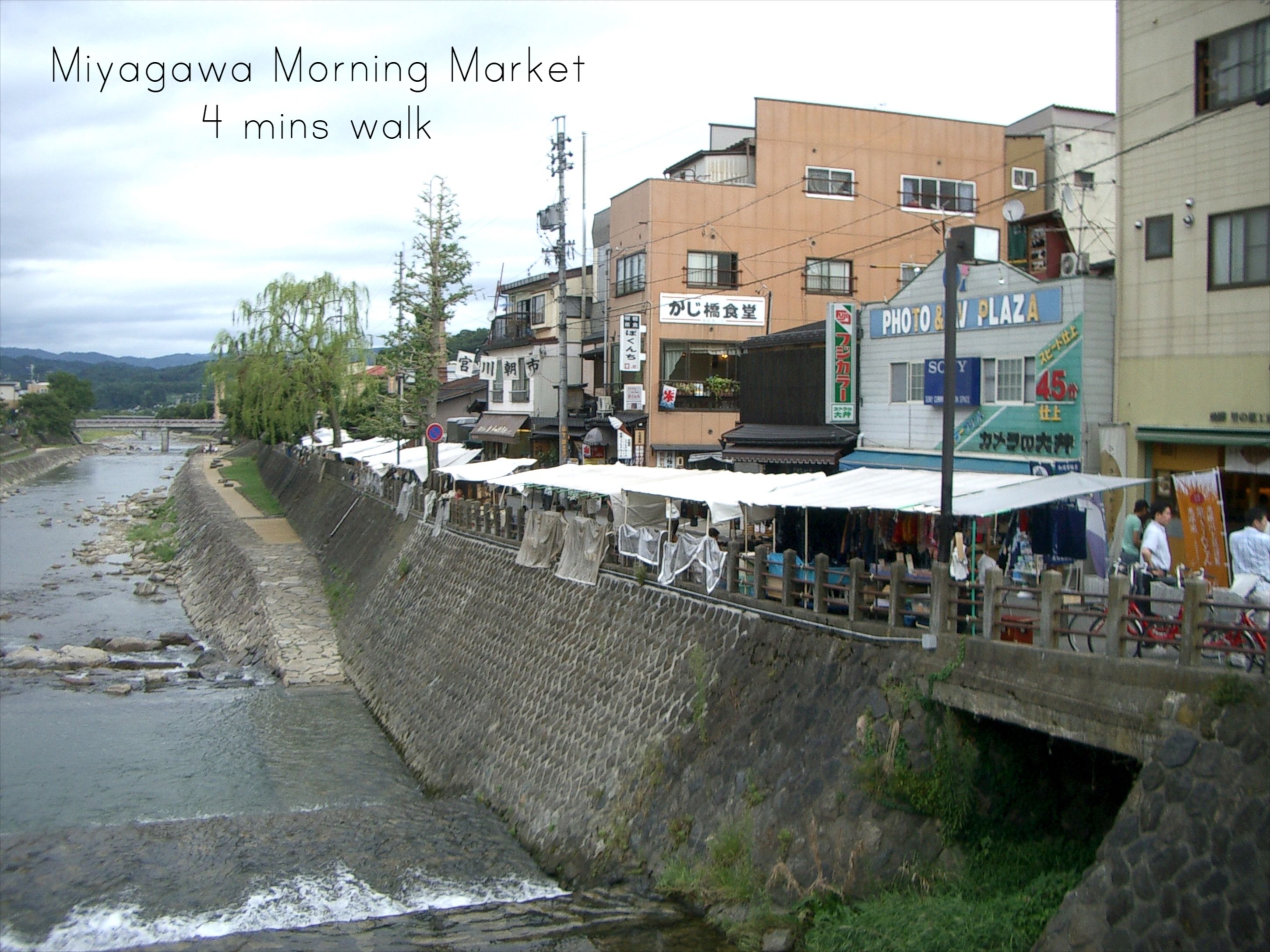 A riverside market with buildings and shops, a stone embankment, and a bridge in the background.