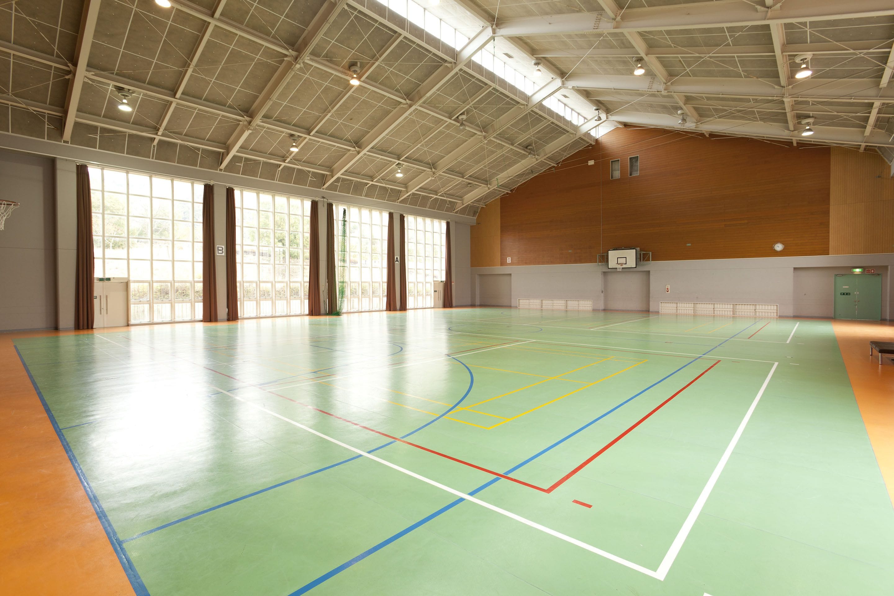 An indoor basketball court with a high ceiling, large windows, and a wooden wall.