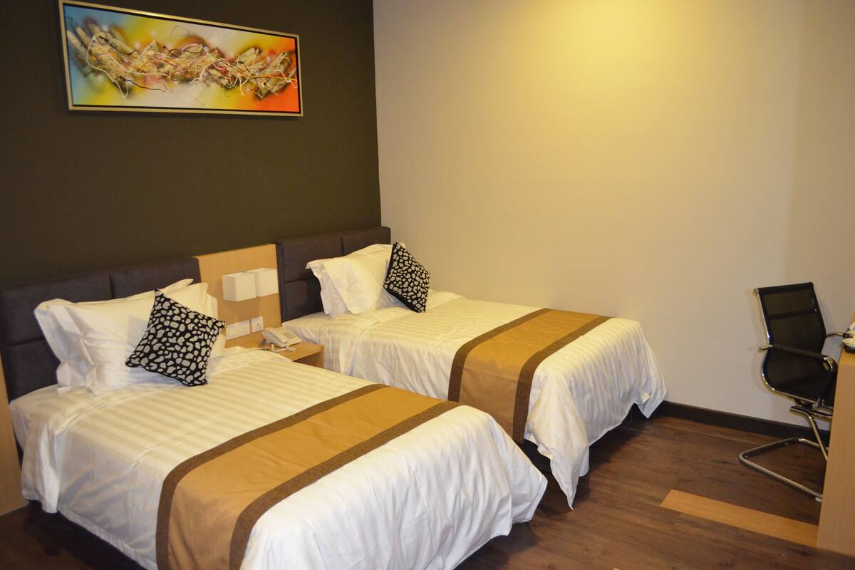 A hotel room with two beds, each with white bedding and brown accents, a bedside table with a phone, a chair, and a painting on the wall.