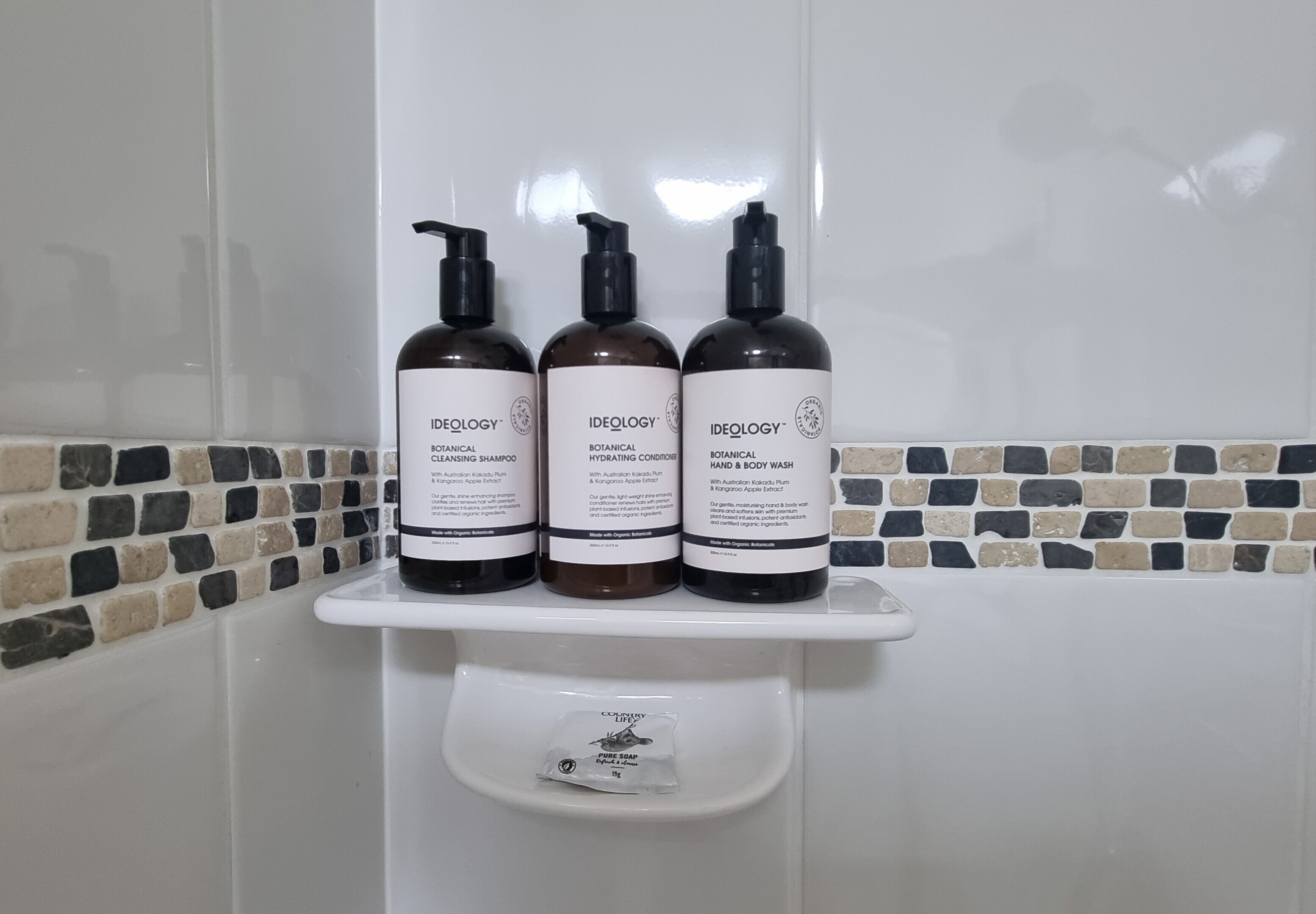 Three bottles of Ideology botanical products on a white shelf in a bathroom with mosaic tile walls.