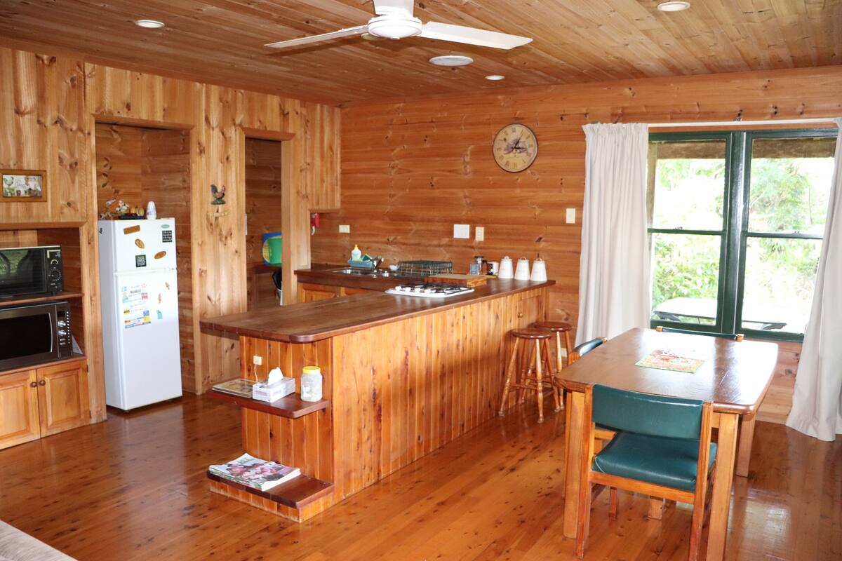A wooden cabin interior with a kitchen, dining area, and a large window.