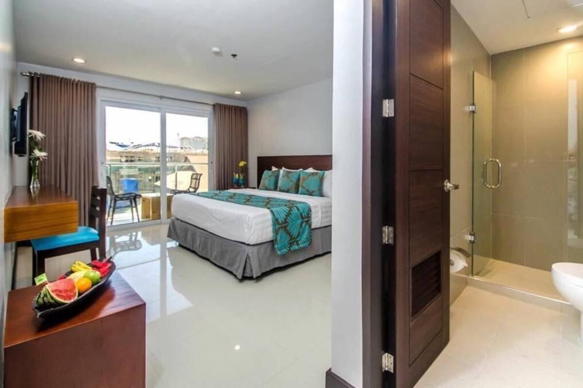 Hotellihuone, jossa on sänky, hedelmillä täytetty pöytä, tuoli, parveke ja suihkutila.