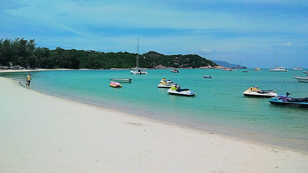 A beach with clear turquoise water, boats, and a person walking along the shore. - Kaya Mani Thai Villa Resort Samui (Koh Samui)
