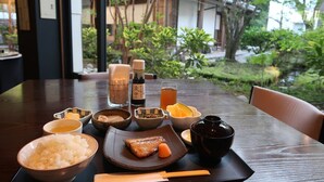Une table à manger dressée avec différents plats, notamment du riz, du poisson et une boisson, le tout mis en valeur par un décor composé d’un bâtiment traditionnel et de verdure.