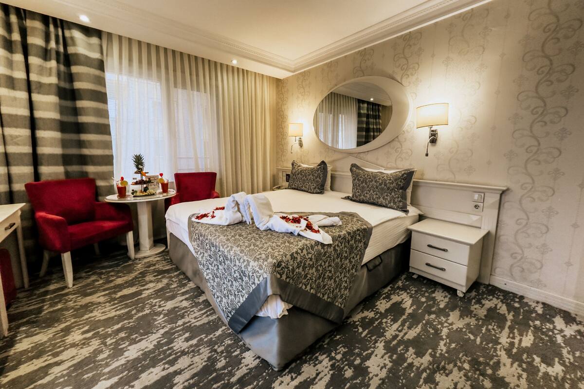 Une chambre d’hôtel avec un grand lit, un fauteuil rouge, une petite table ronde avec des boissons et des en-cas, un miroir et une moquette à motifs.