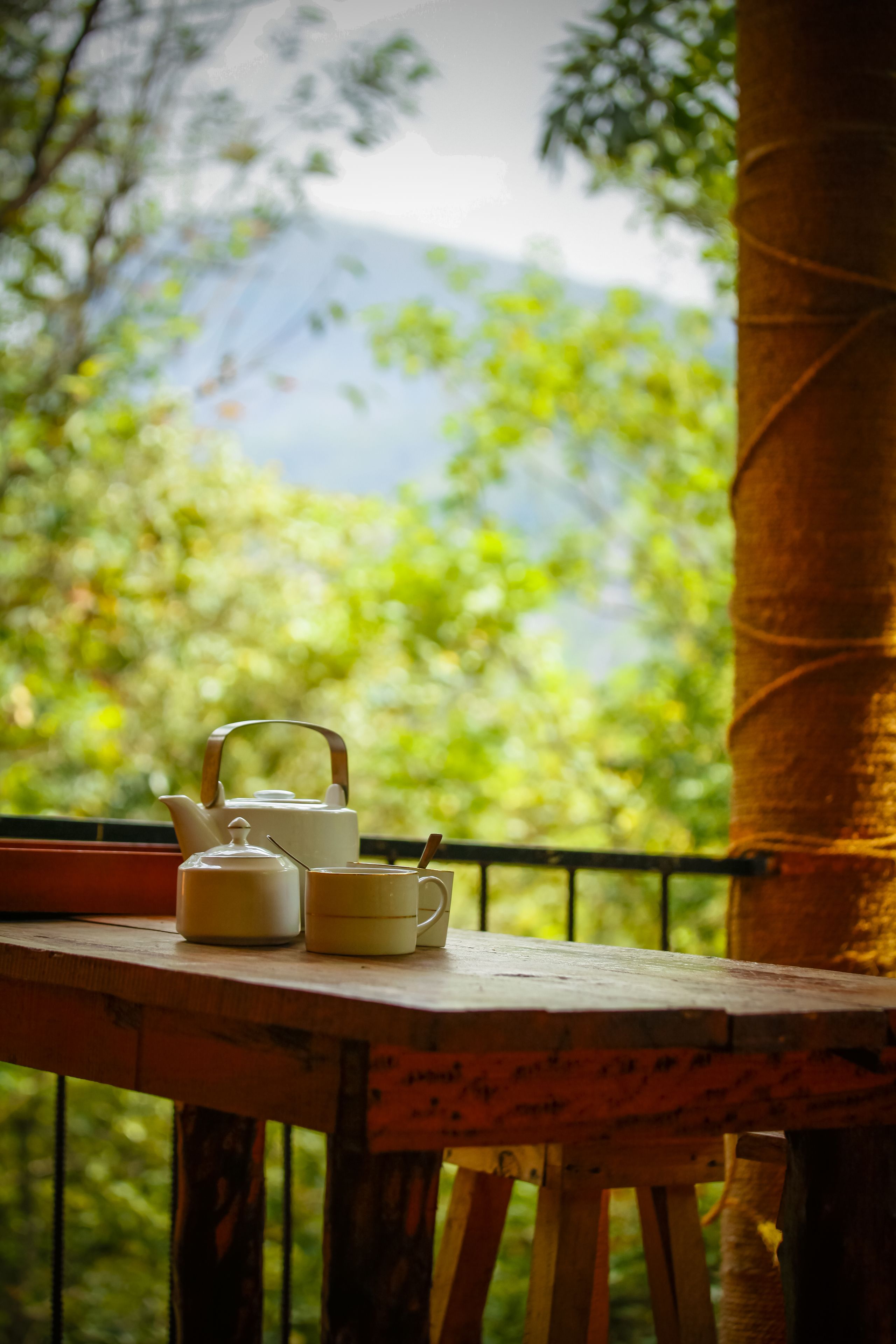 A wooden table with a teapot, cup, and saucer on a balcony with a view of trees and mountains.