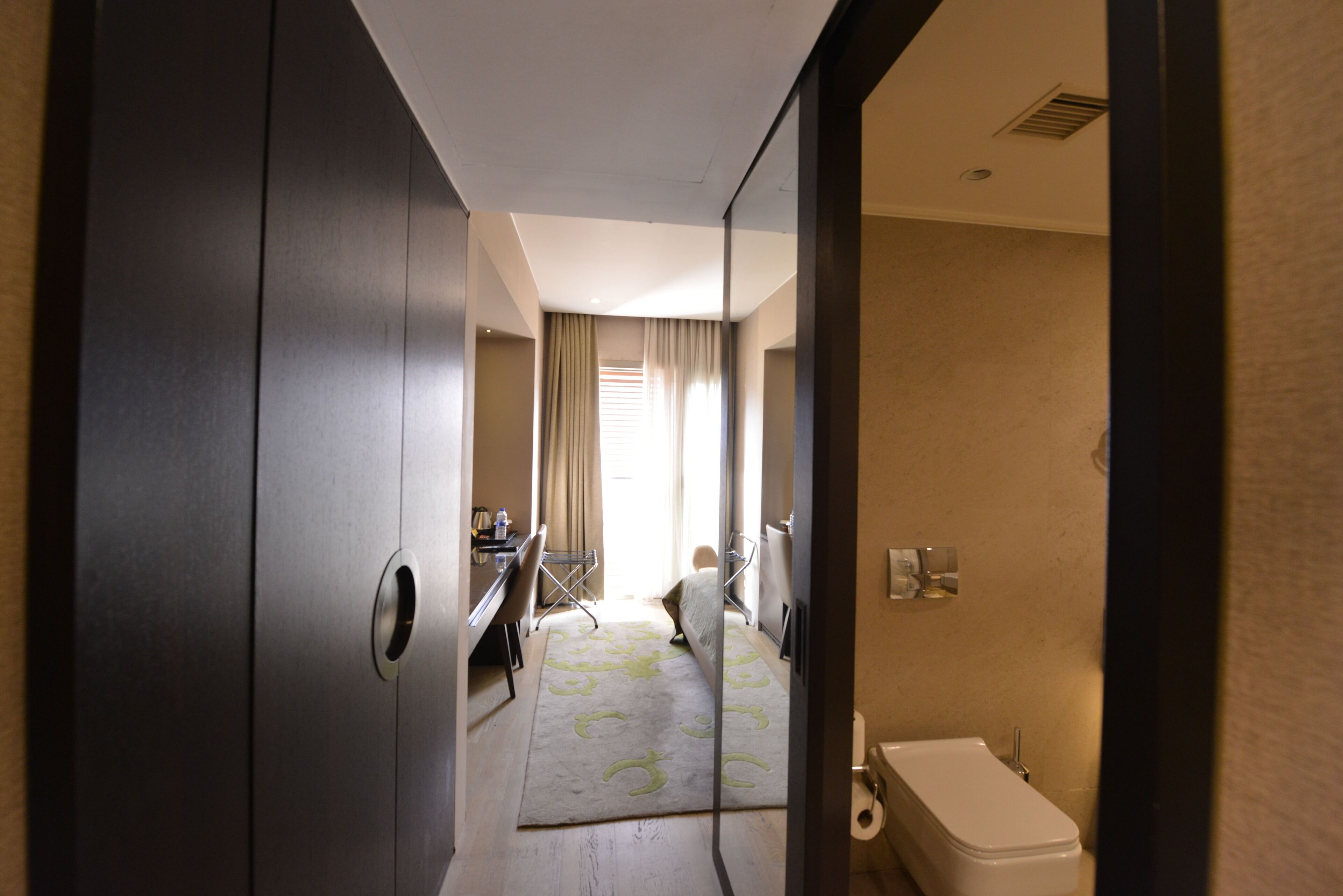 Habitación de hotel con un armario grande, un escritorio con silla, una cama y un baño con inodoro y lavamanos.