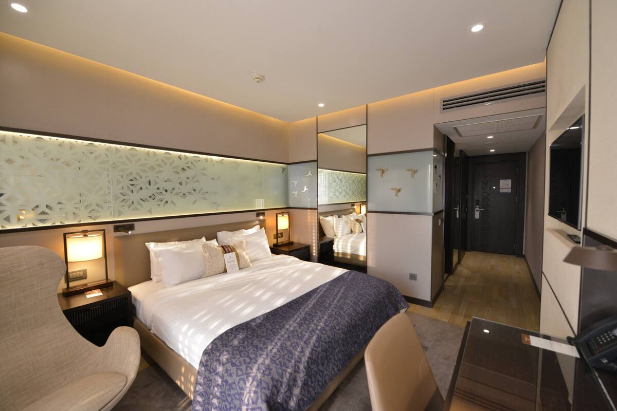 A modern hotel room with a large bed, bedside tables with lamps, a seating area with a chair, and a mirror.