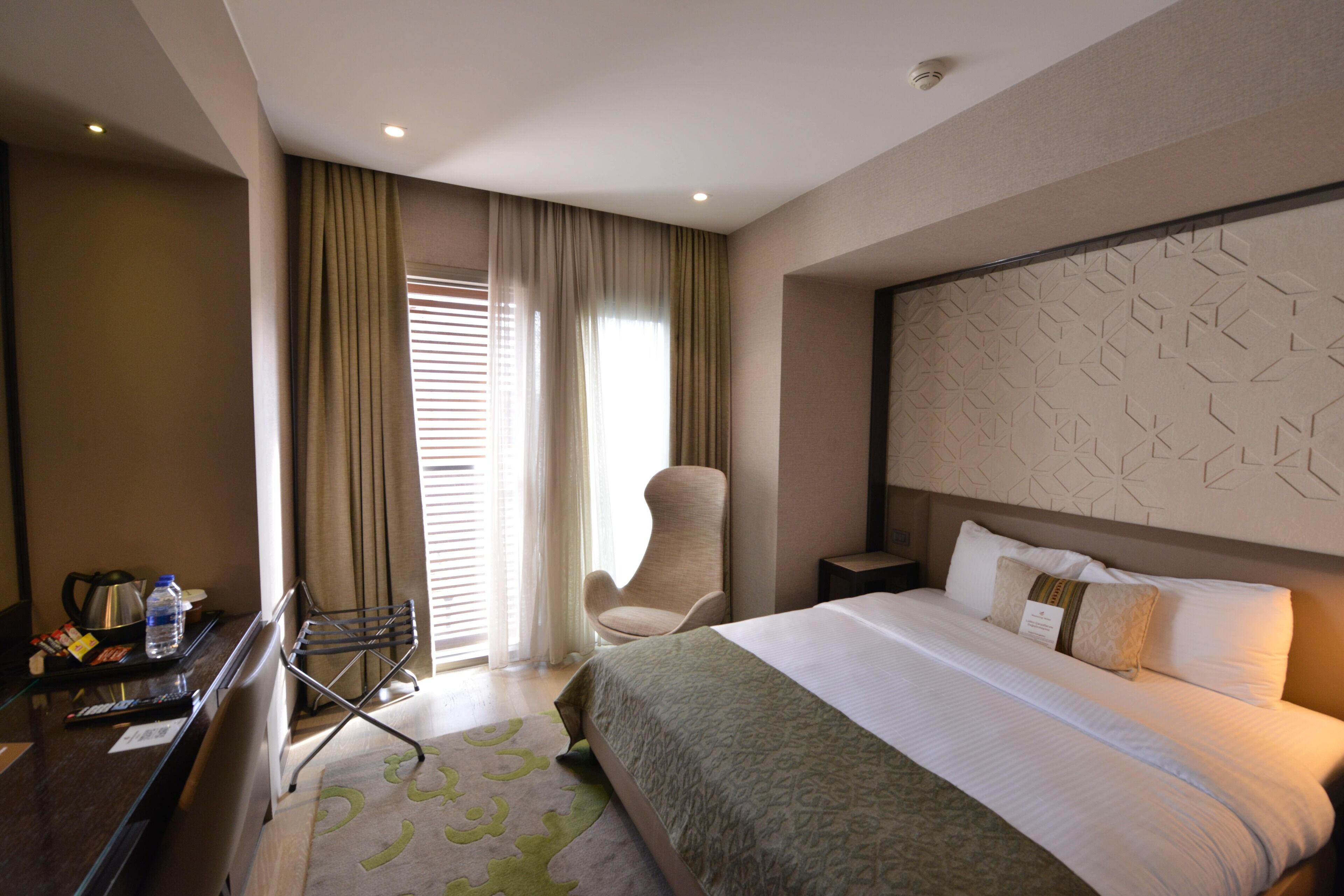 A hotel room with a bed, a chair, a desk with a kettle, and a window with curtains.