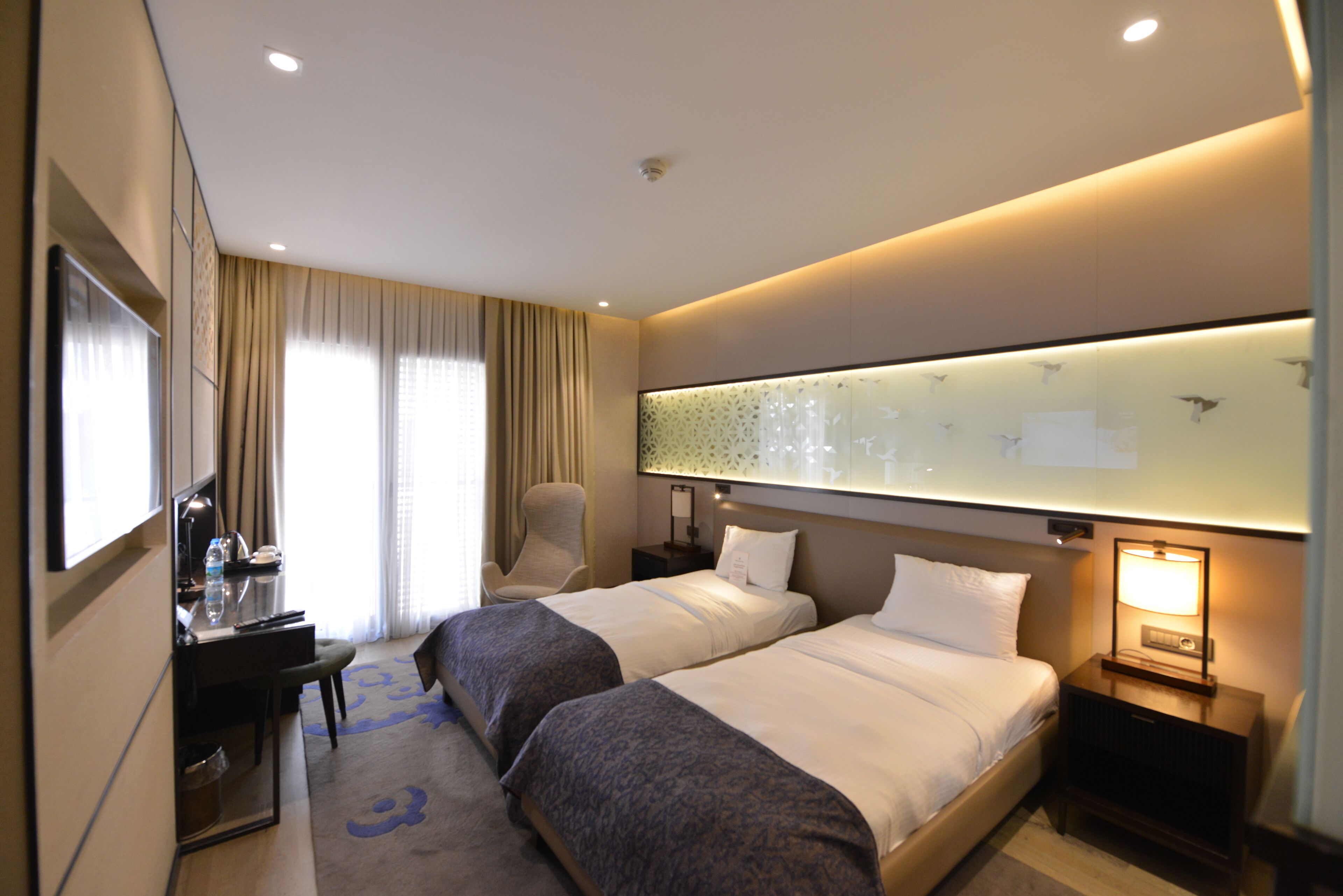 A hotel room with two beds, a desk, a chair, a mirror, and a nightstand with a lamp.