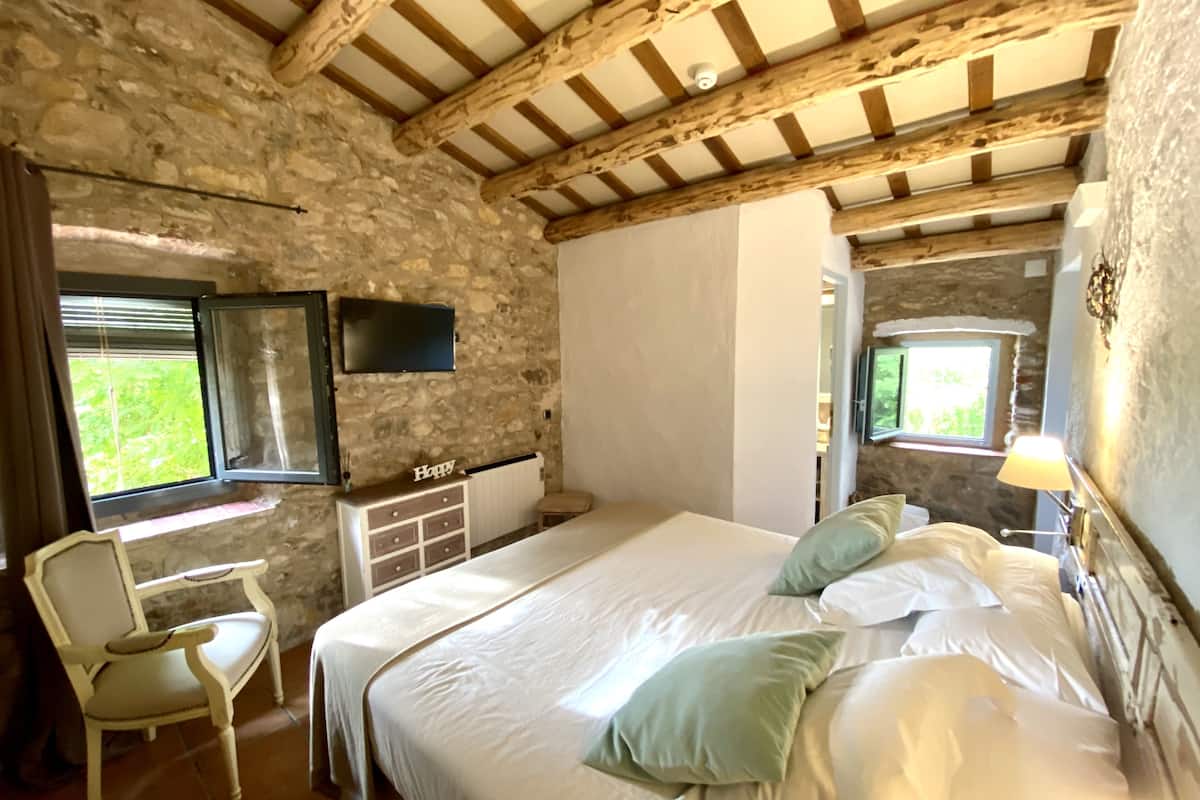 Een slaapkamer met een stenen muur, houten balken, een bed met wit beddengoed, een stoel en een televisie aan de muur.