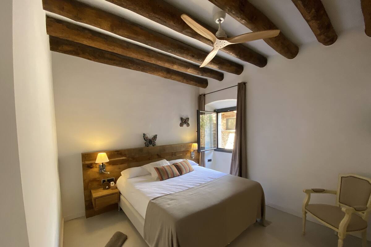 Een slaapkamer met een houten plafond, een bed met een beige dekbedovertrek, een nachtkastje met een lamp, een stoel en een raam met gordijnen.