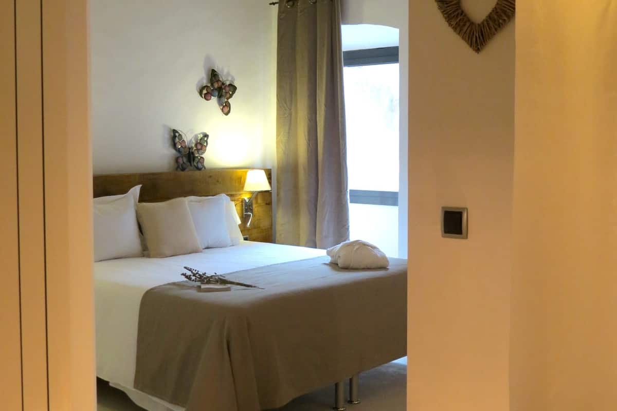 Een slaapkamer met een bed, een nachtkastje, een lamp, een raam met gordijnen en een hartvormige decoratie aan de muur.