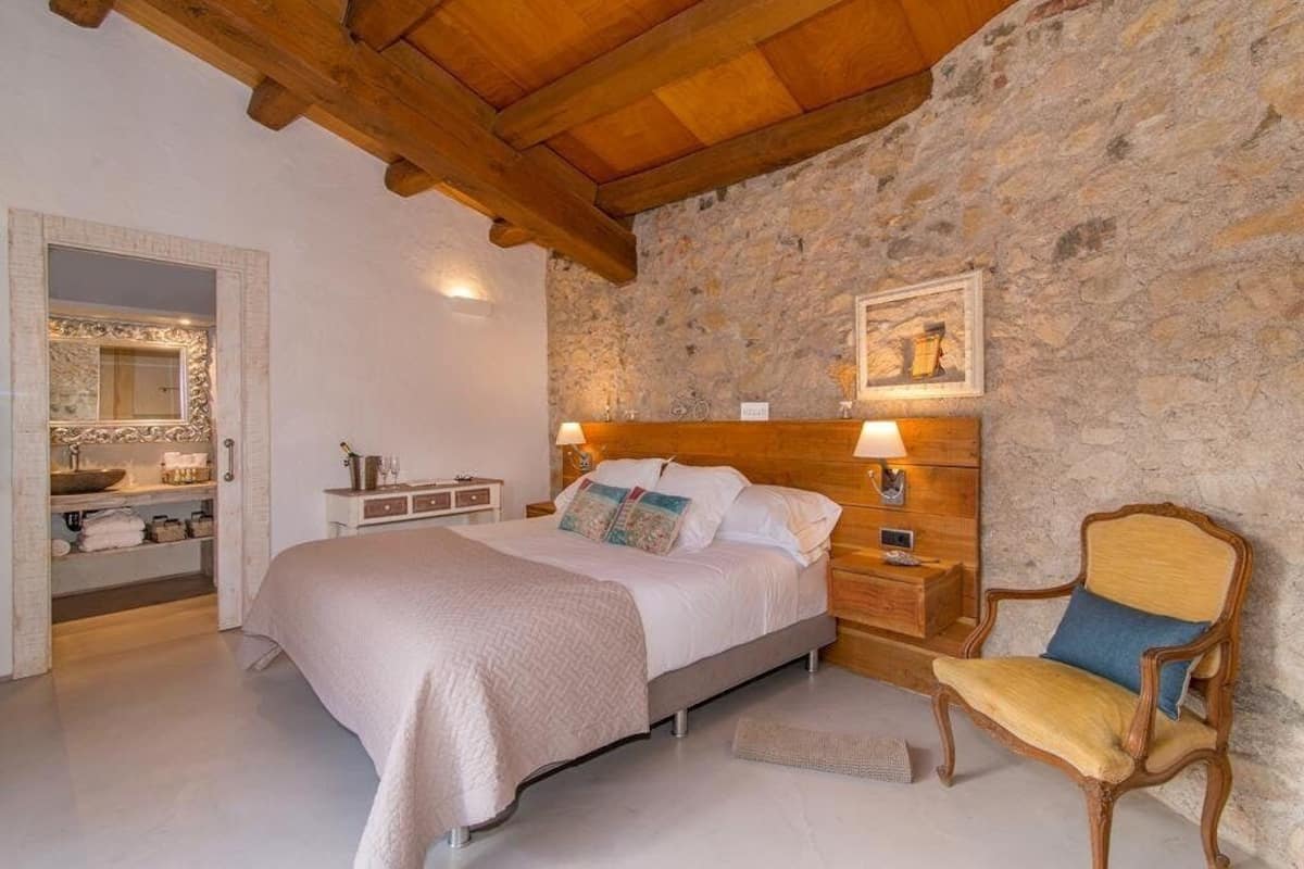 Een slaapkamer met een stenen muur, houten balken in het plafond, een bed met een witte dekbedovertrek, een nachtkastje met een lamp en een stoel.