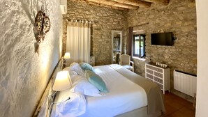A rustic room with a stone wall, a bed with white linens, a lamp, a chair, a dresser, and a television mounted on the wall.