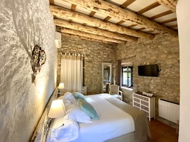 Una habitación rústica con pared de piedra, una cama con ropa blanca, una lámpara, una silla, un tocador y un televisor montado en la pared.