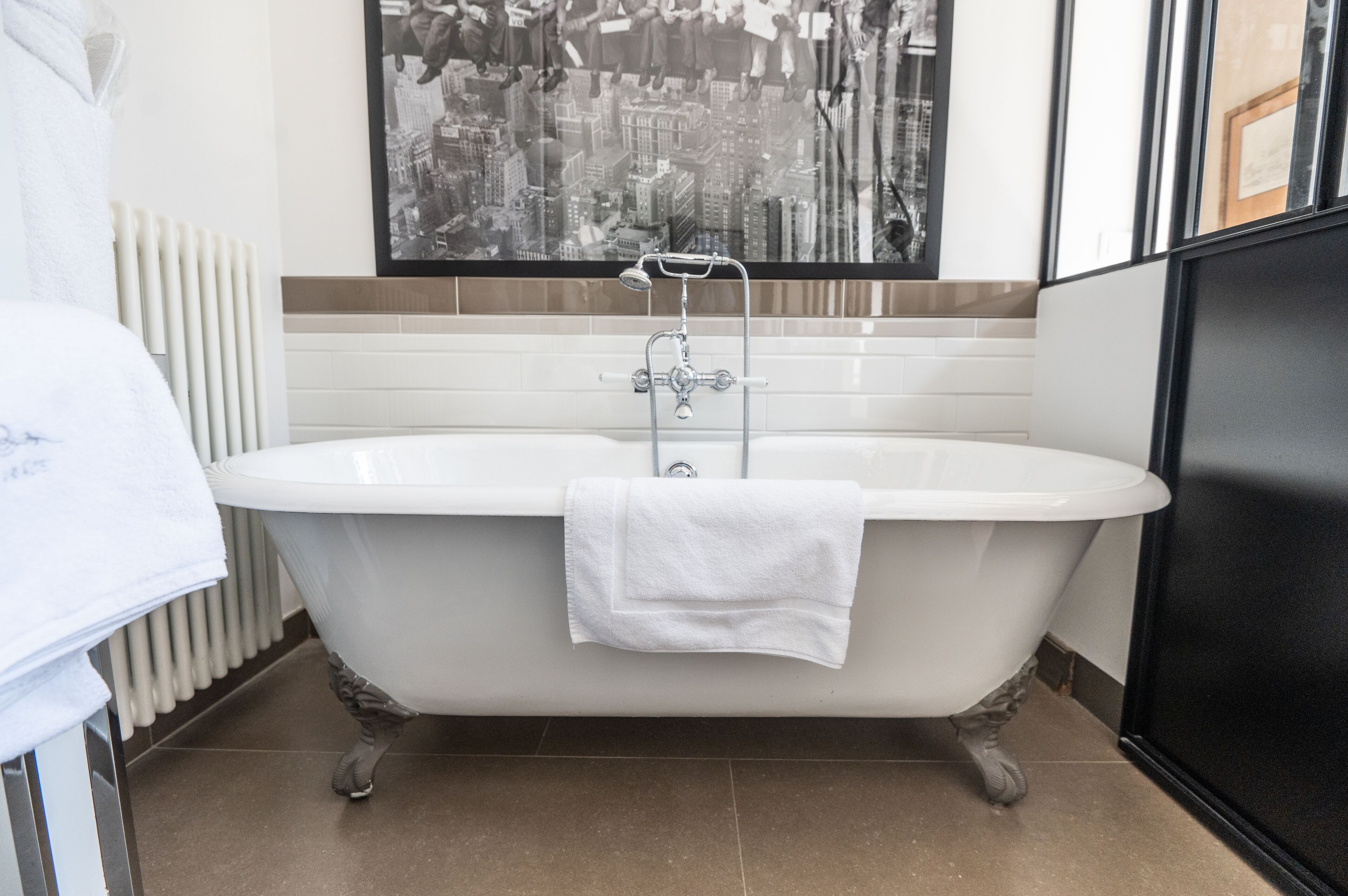 Ett badrum med ett fristående badkar, en vit handduk över kanten och ett konstverk med en stadsbild på väggen.