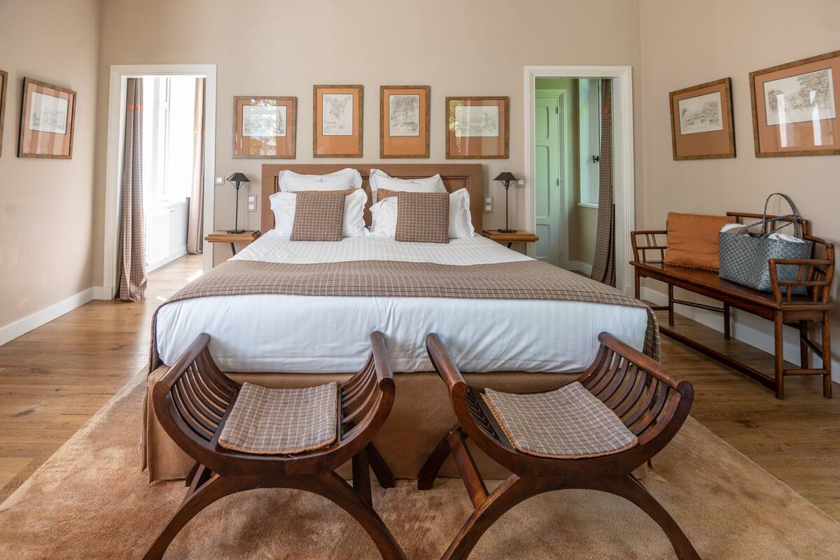 Um quarto espaçoso com uma cama grande, duas cadeiras de madeira, uma mesinha e um banco.
