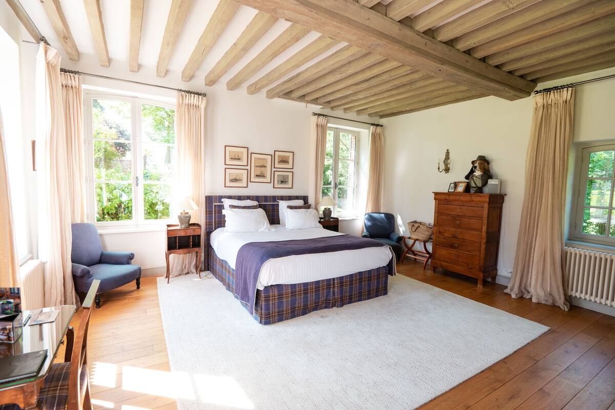 Quarto com cama grande, vigas de madeira, cômoda, cadeira e janela com cortinas.