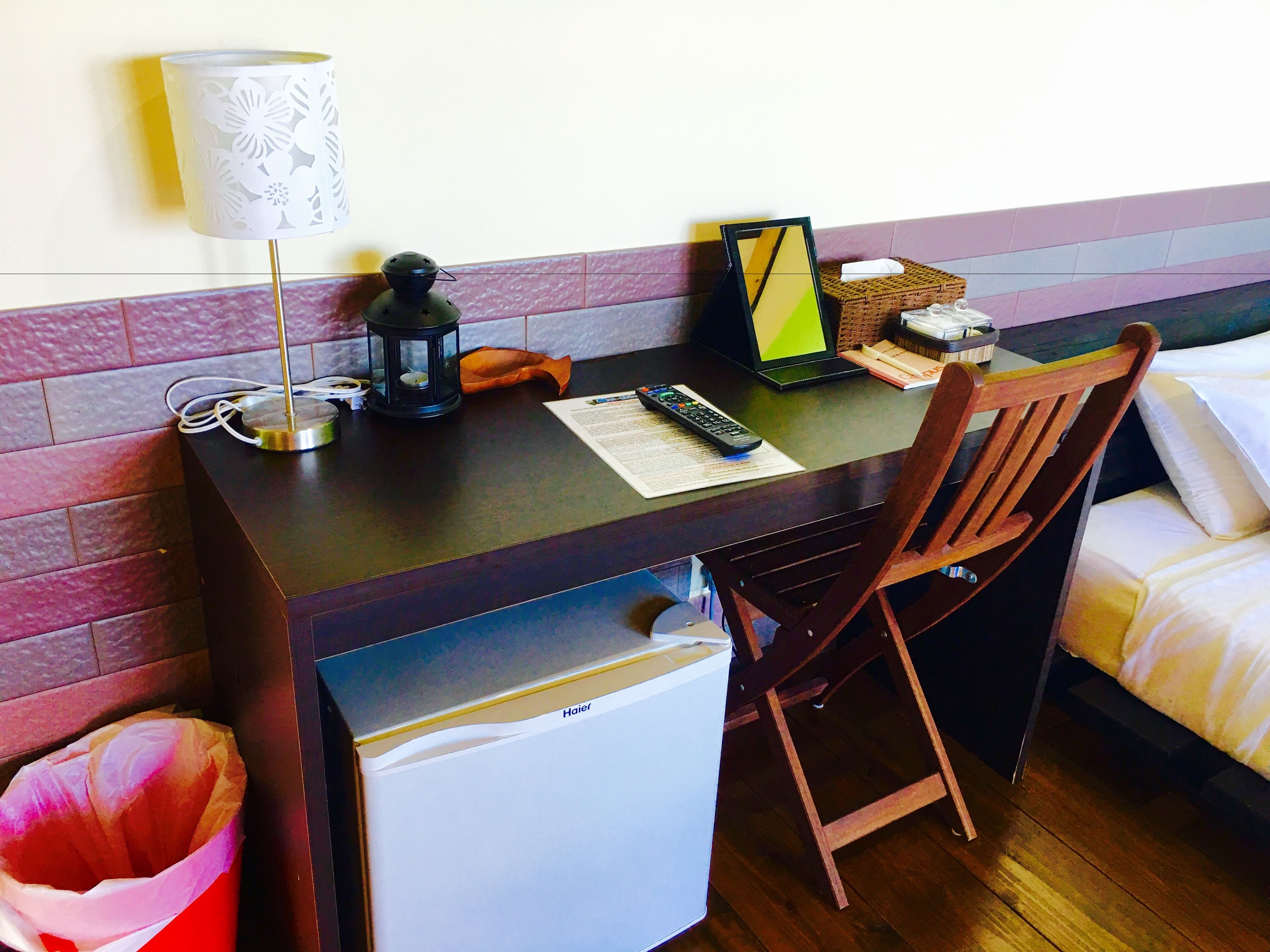 Hotelkamer met een houten bureau, een stoel, een kleine koelkast, een lamp, een lantaarn, een bed en een nachtkastje met een zakdoosje.