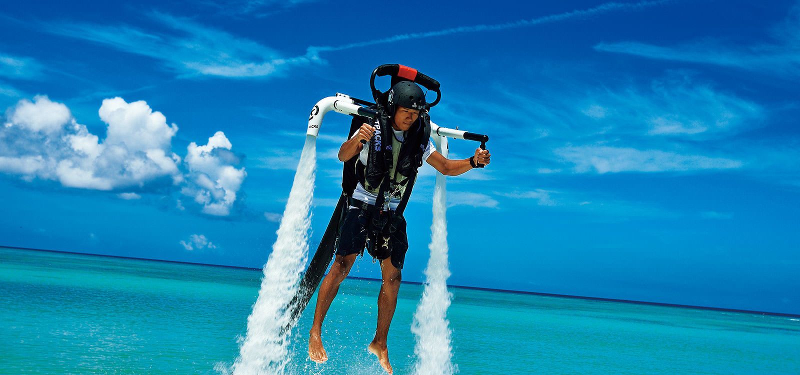 A person water skiing with a jet ski, wearing a helmet and life vest, against a clear blue sky and ocean backdrop.