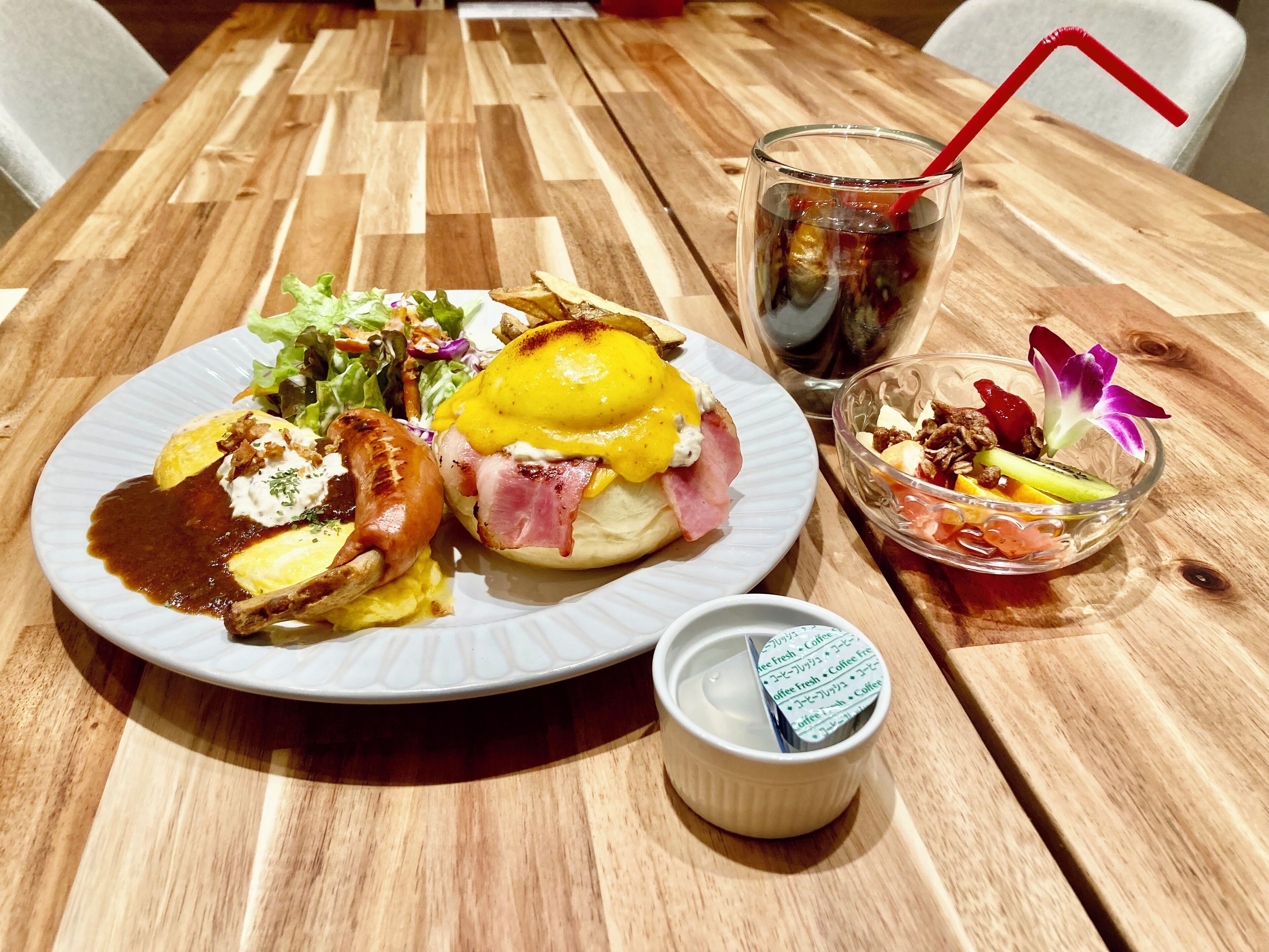A wooden table with a plate of food, a glass of iced coffee, and a small bowl of fruit.