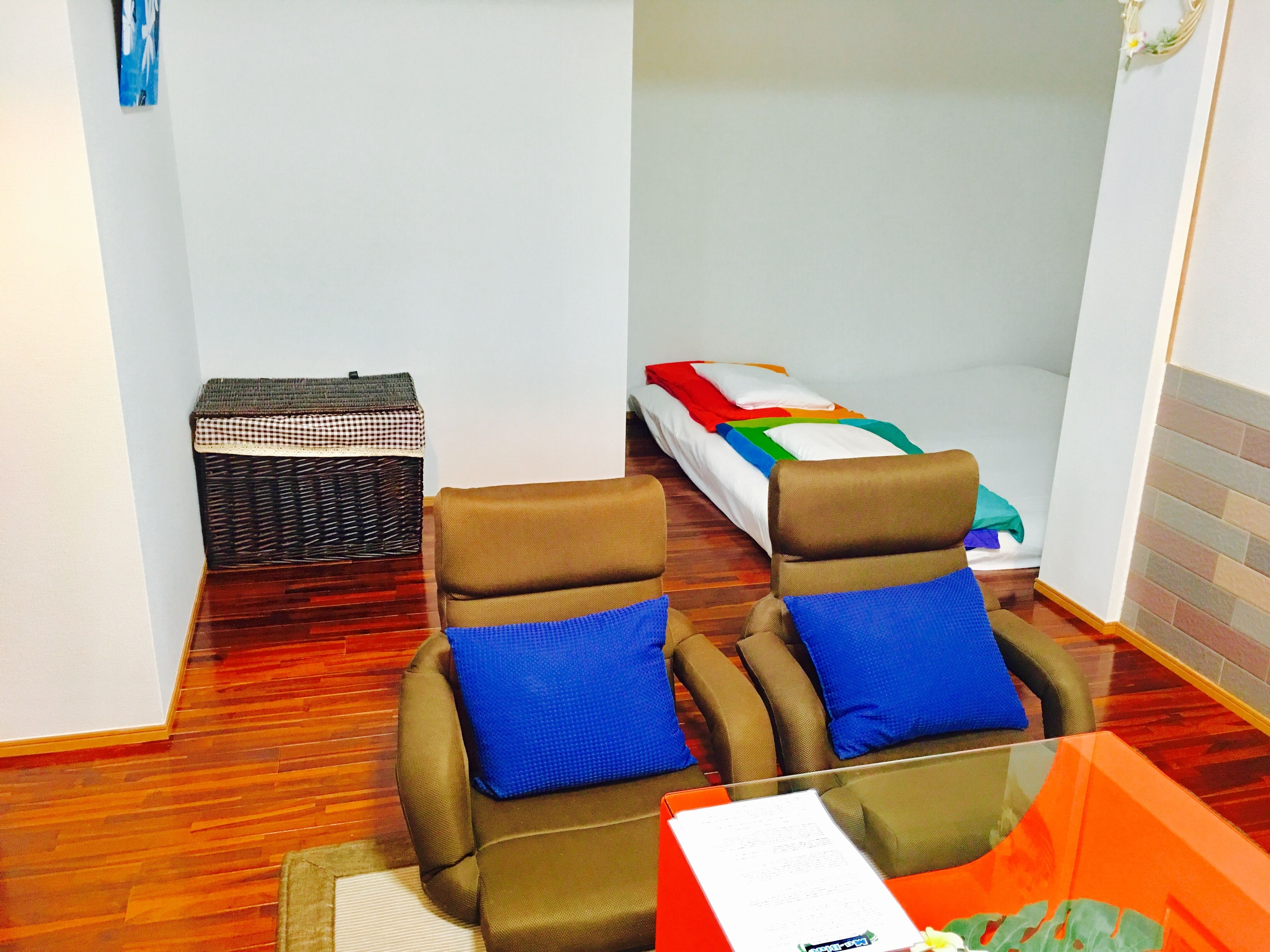 A room with two brown armchairs with blue cushions, a small table with a menu, a wicker basket, and a bed with colorful bedding.