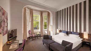 Een hotelkamer met een groot bed, twee nachtkastjes met lampen, een stoel, een klein tafeltje met een schaal fruit, en een groot raam met gordijnen.