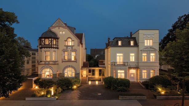 Een historisch gebouw met een toren en erkers, omringd door een fraai aangelegde tuin en een geplaveid pad.