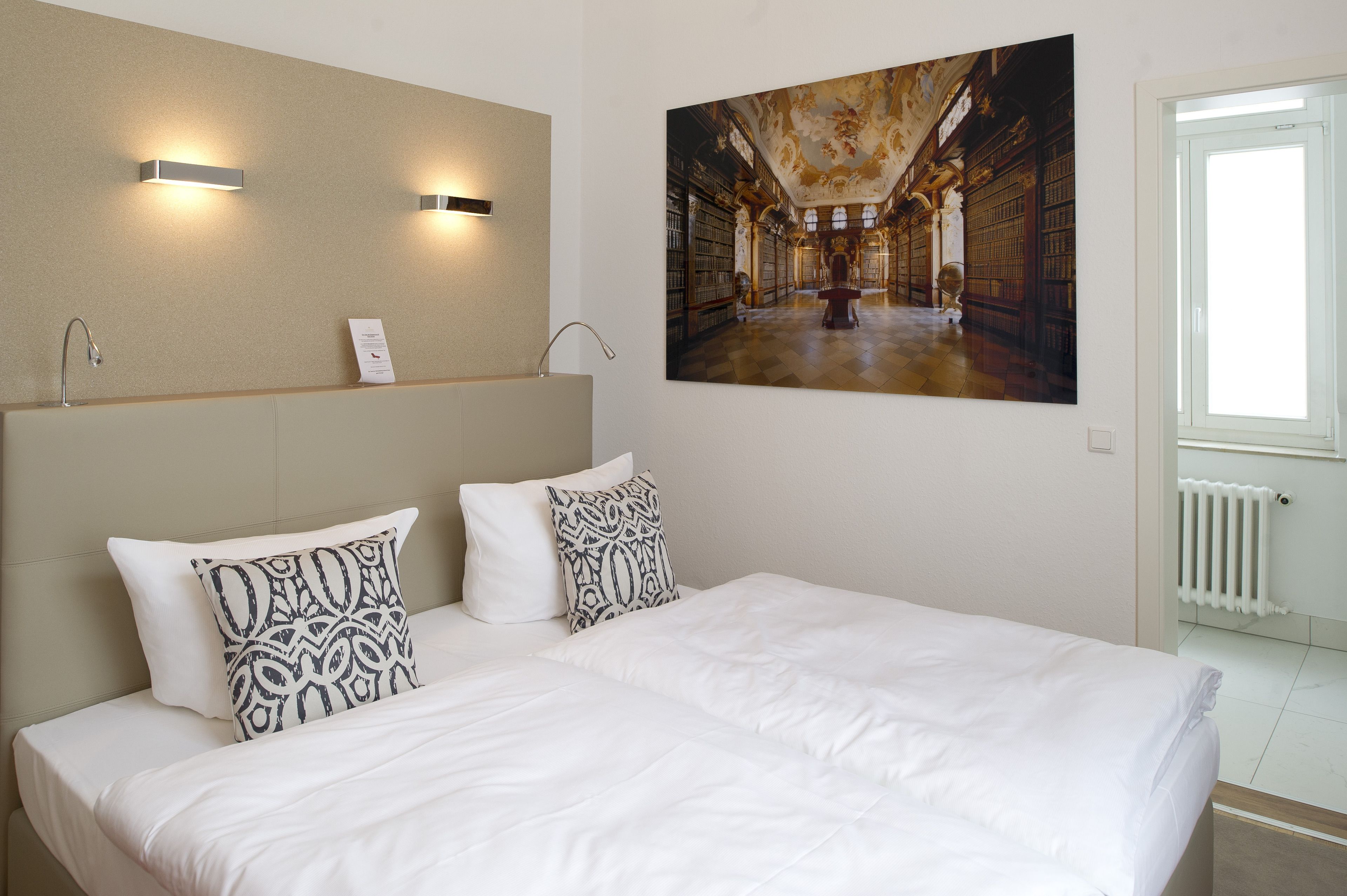 Une chambre d’hôtel avec un grand lit, deux appliques murales, un tableau représentant une bibliothèque et une fenêtre avec un radiateur.