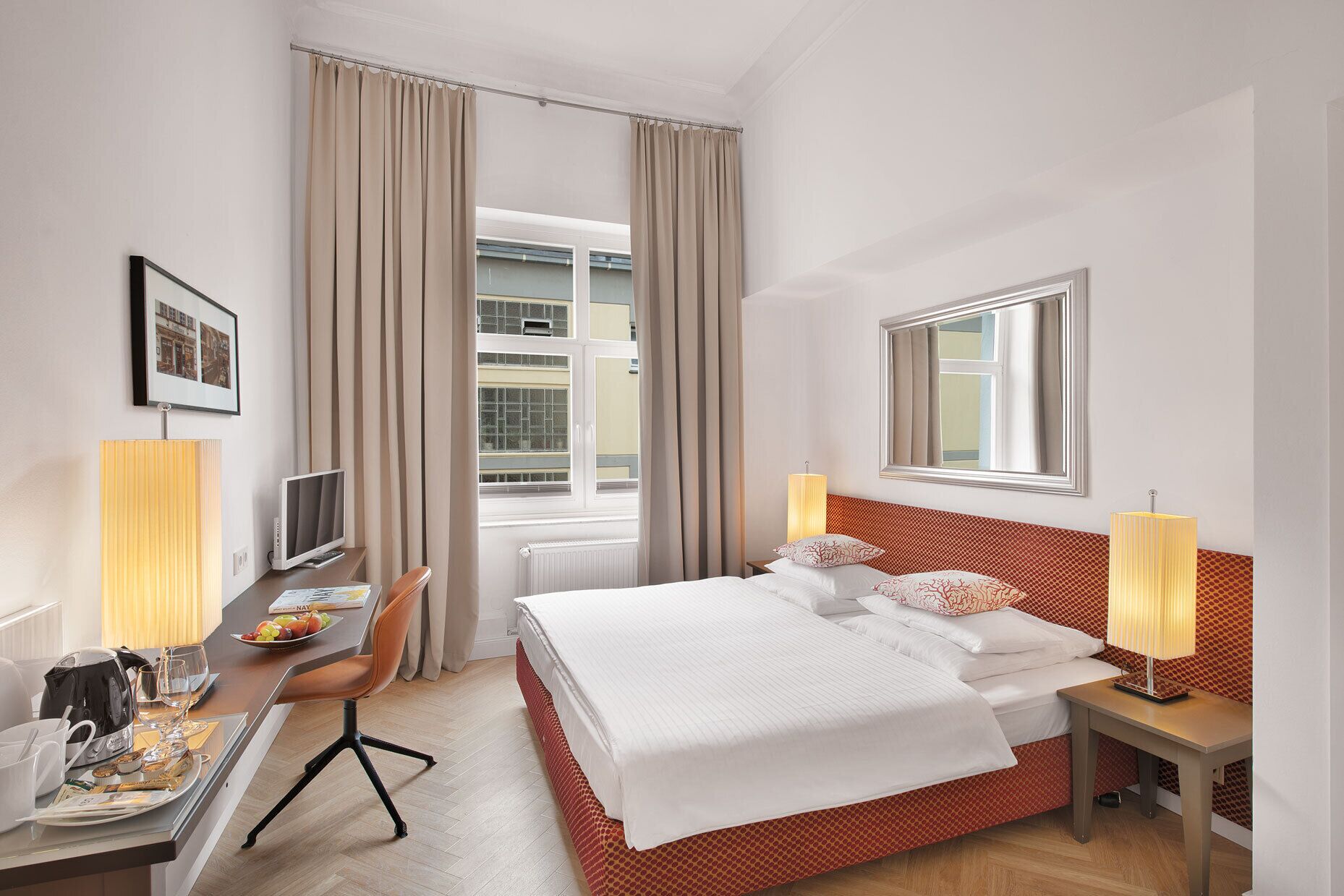 A hotel room with a bed, a desk with a computer, a chair, a window with curtains, and a lamp.