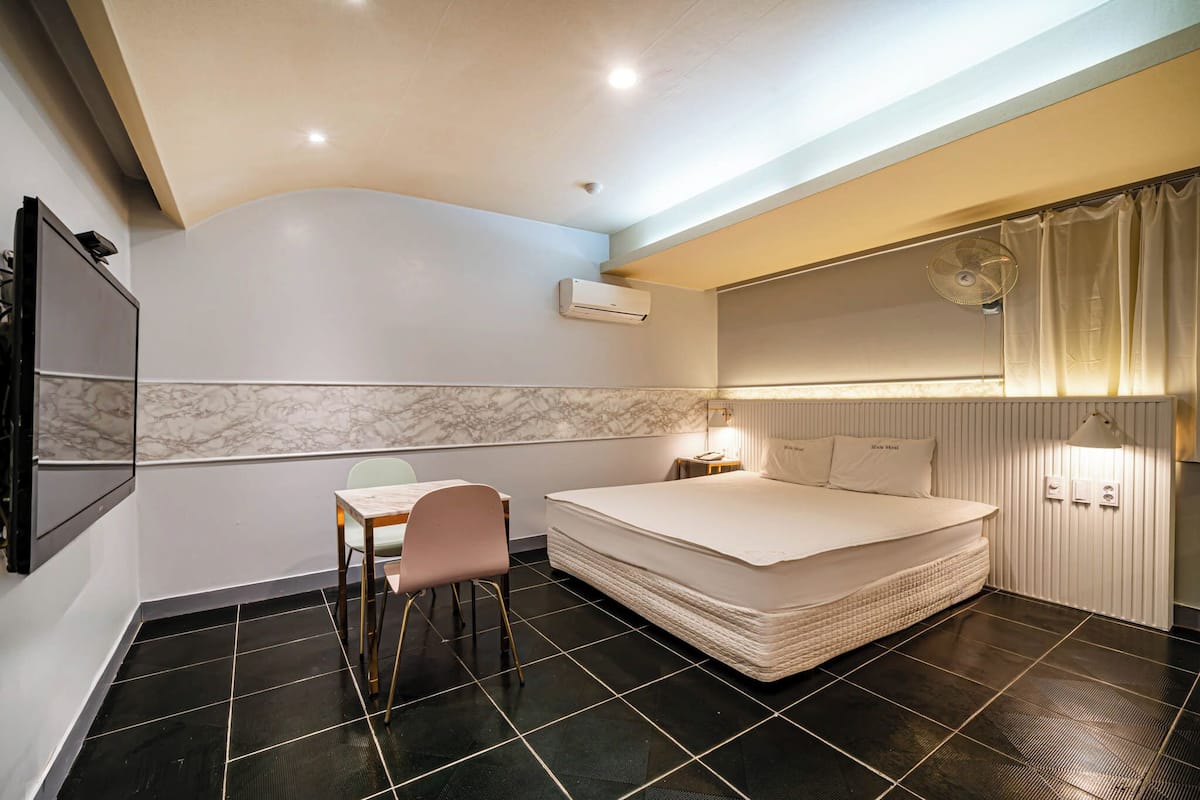 A bedroom with a bed, a desk, a chair, an air conditioning unit, and a wall-mounted TV.
