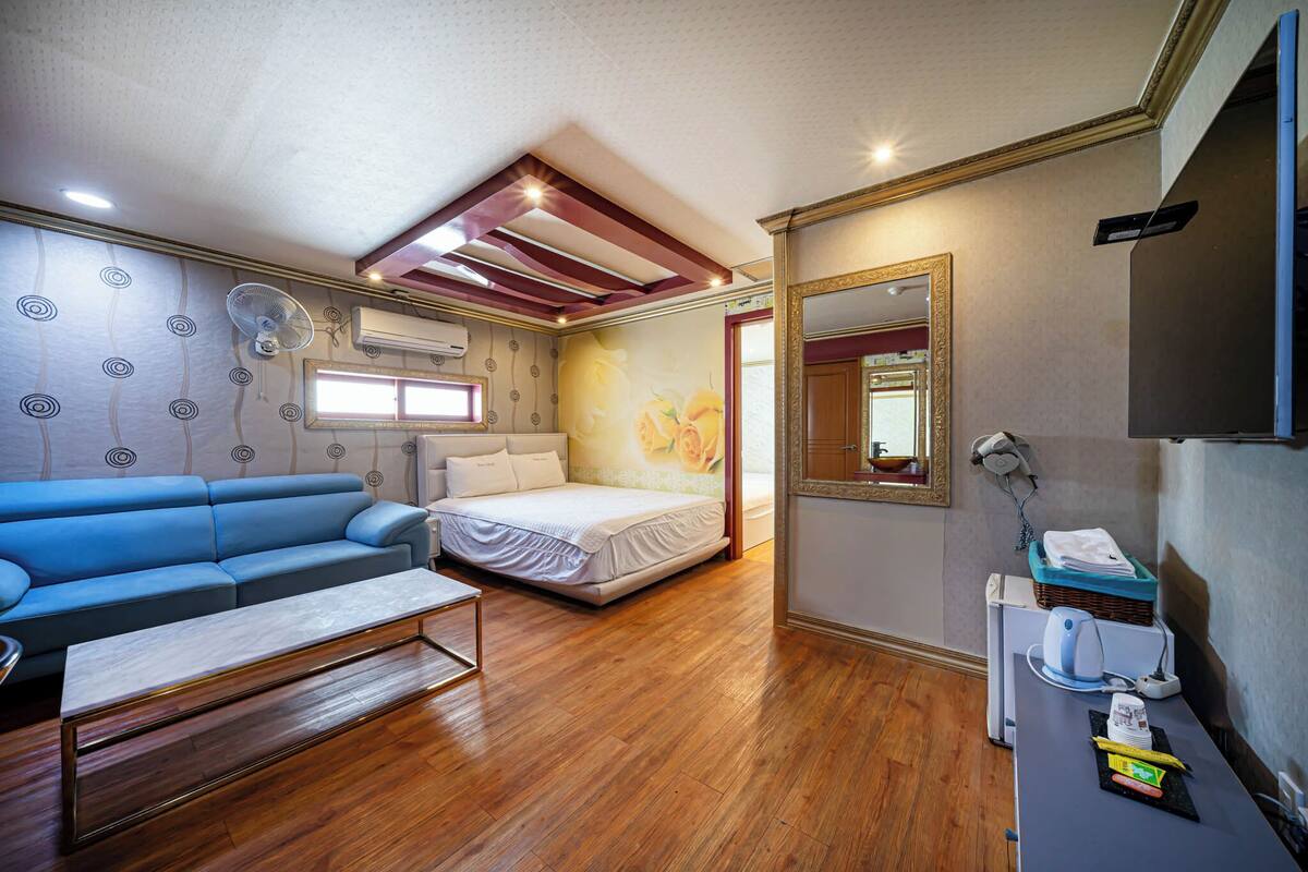 A compact room with a blue sofa, a single bed, a wooden floor, a ceiling fan, and a mirror.
