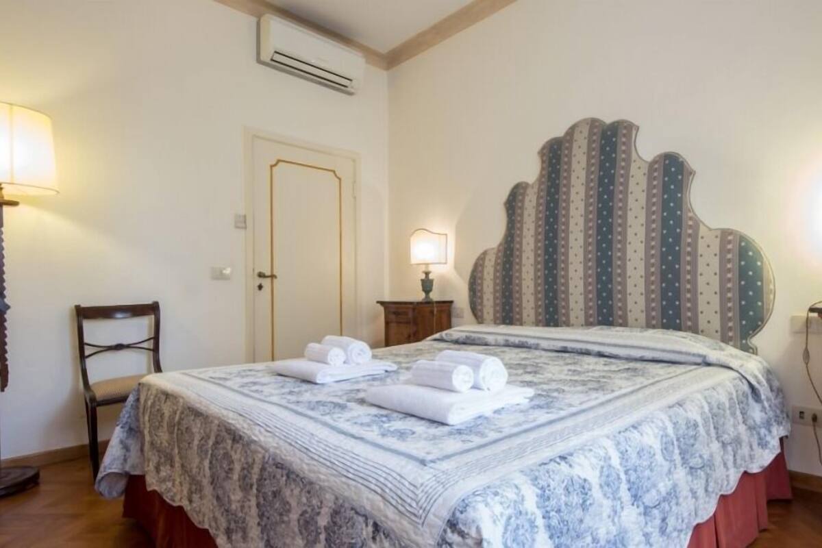 A neatly made bed with a patterned bedspread and two rolled towels, a wooden chair, a bedside table with a lamp, and a door in a hotel room.
