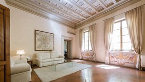 A spacious room with a high ceiling, a large window with curtains, a sofa, a small table with a lamp, and a framed map on the wall. - Florentapartments - Ponte Vecchio (Florence)