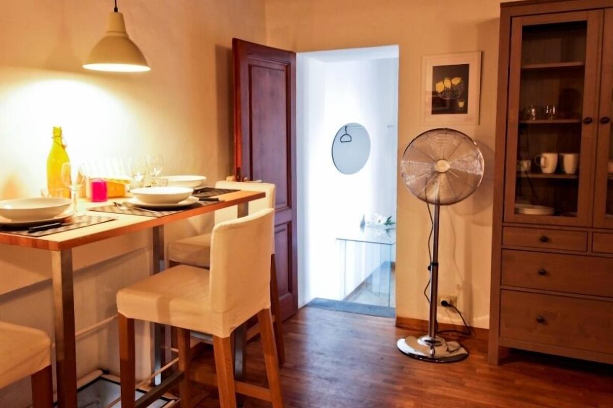A dining area with a wooden table, white chairs, a bottle, glasses, and a fan.