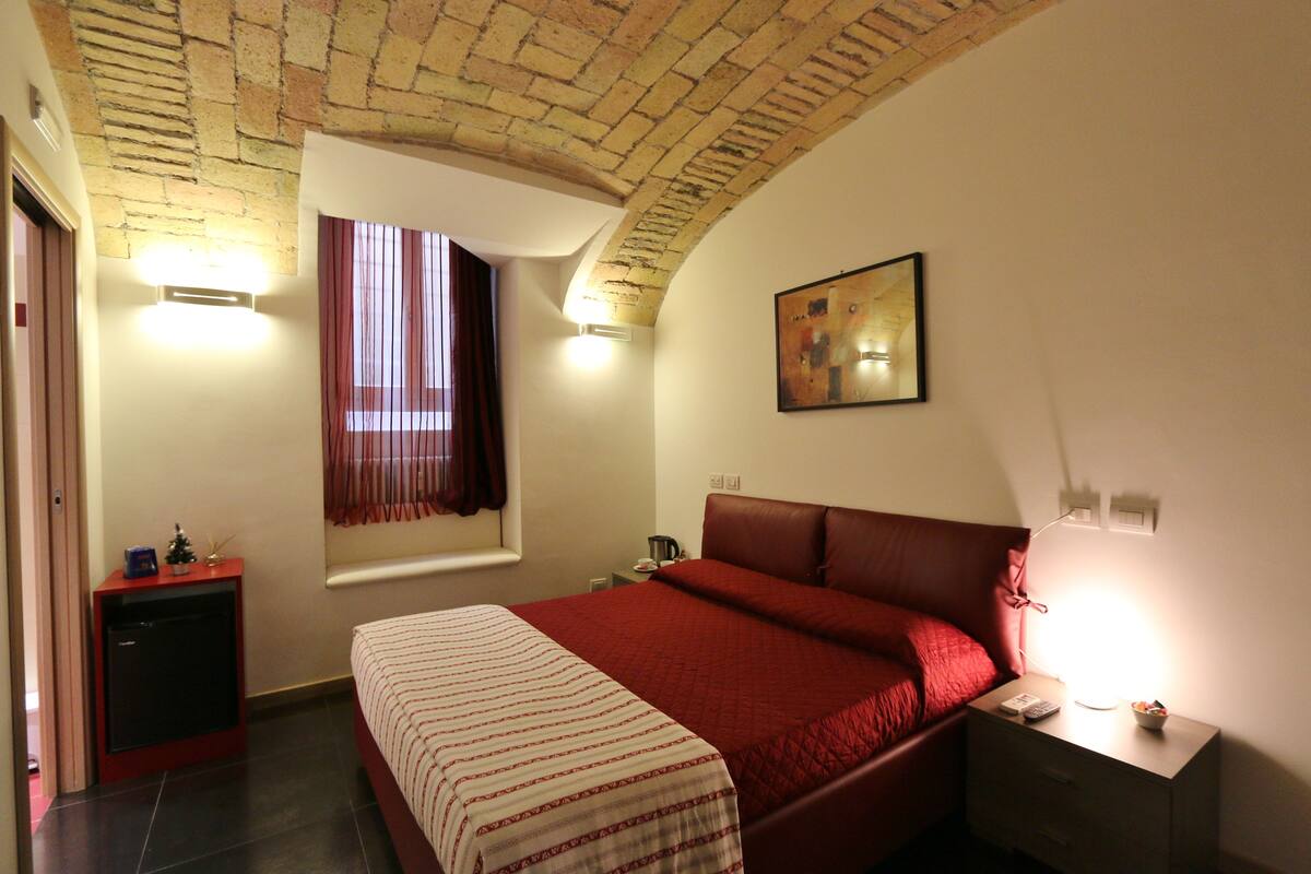 A bedroom with a brick ceiling, a bed with a red bedspread, a small bedside table with a lamp, and a window with curtains.